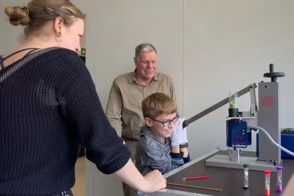 Ein Schüler mit Brille und Handschuhen führt ein Experiment an einer Maschine namens „HollMaker“ durch, während zwei Erwachsene aufmerksam zuschauen – Teil des SKZ-Schülerlabors „Plastik verstehen – Zukunft gestalten“.