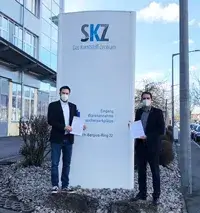 Zwei Vertreter des SKZ, Dr. Achenbach und Dr. Baudrit, stehen vor dem Eingangsschild des Kunststoff-Zentrums und halten Dokumente in den Händen – ein Zeichen der Unterstützung durch die STAHLGRUBER GESELLSCHAFTER-STIFTUNG.