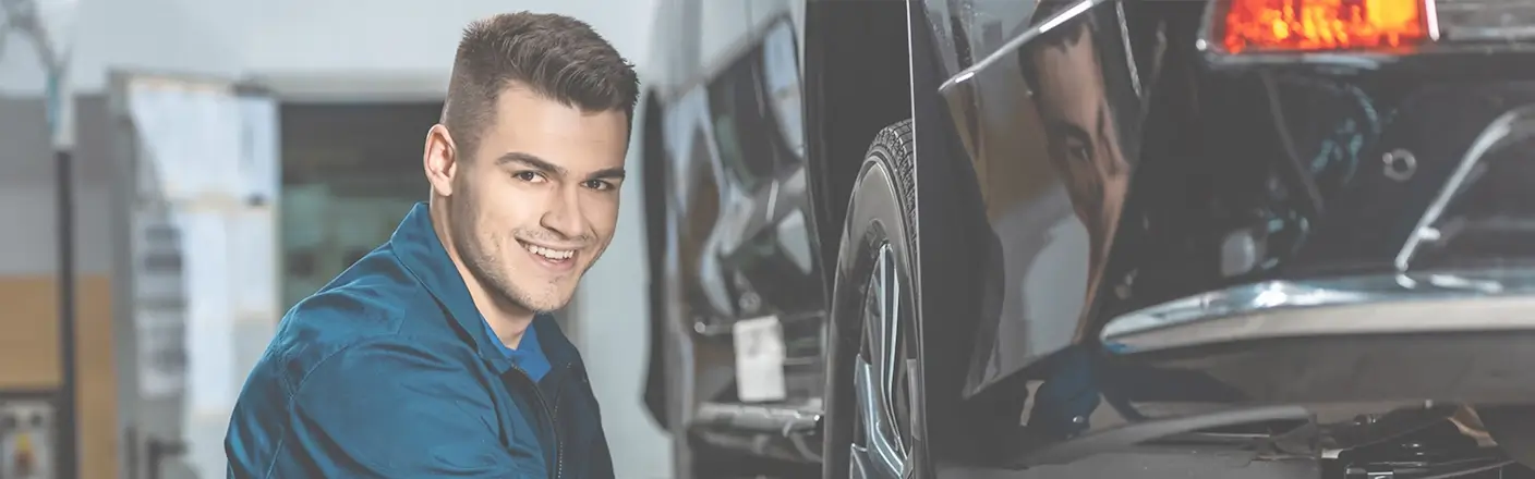 Lächelnder Automechaniker in blauer Arbeitskleidung neben einem Auto in einer Werkstatt